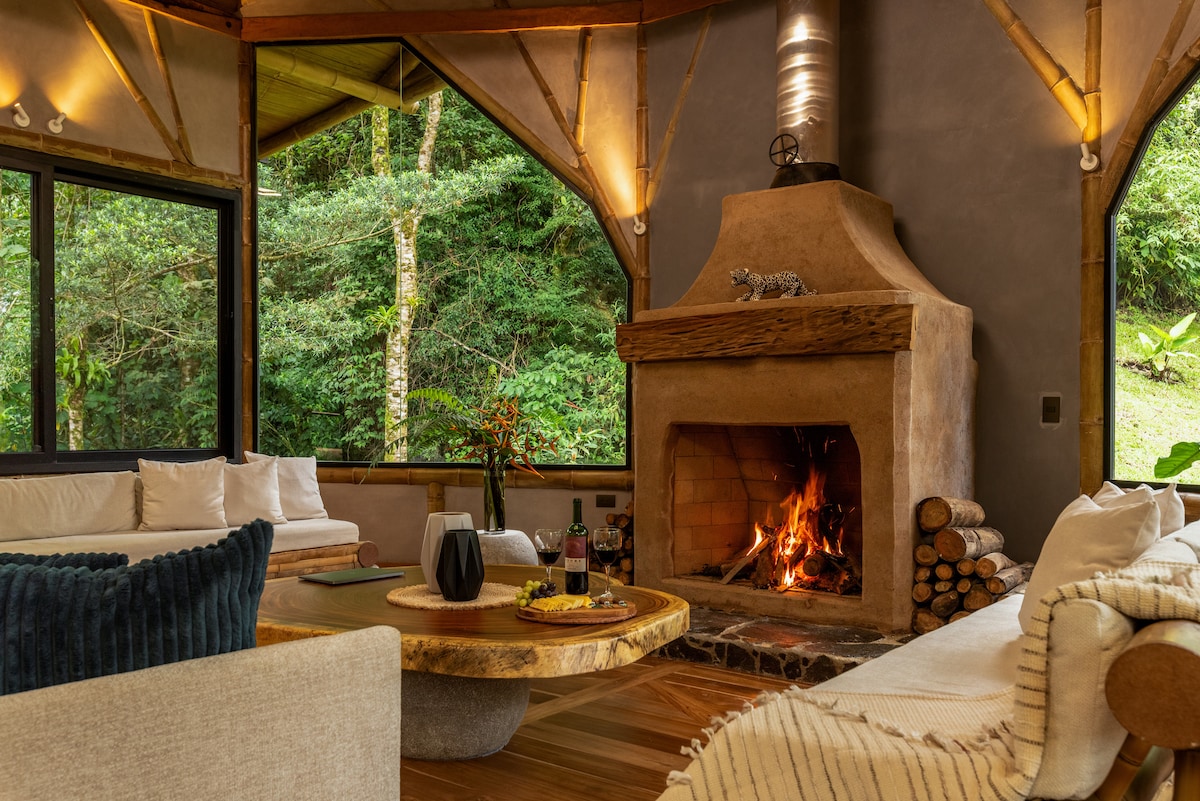A cozy living area features a rustic stone fireplace, surrounded by comfortable seating arrangements. Large windows provide views of the lush greenery outside, while a wooden coffee table sits in the center, adorned with snacks and a bottle of wine, welcoming moments of relaxation.