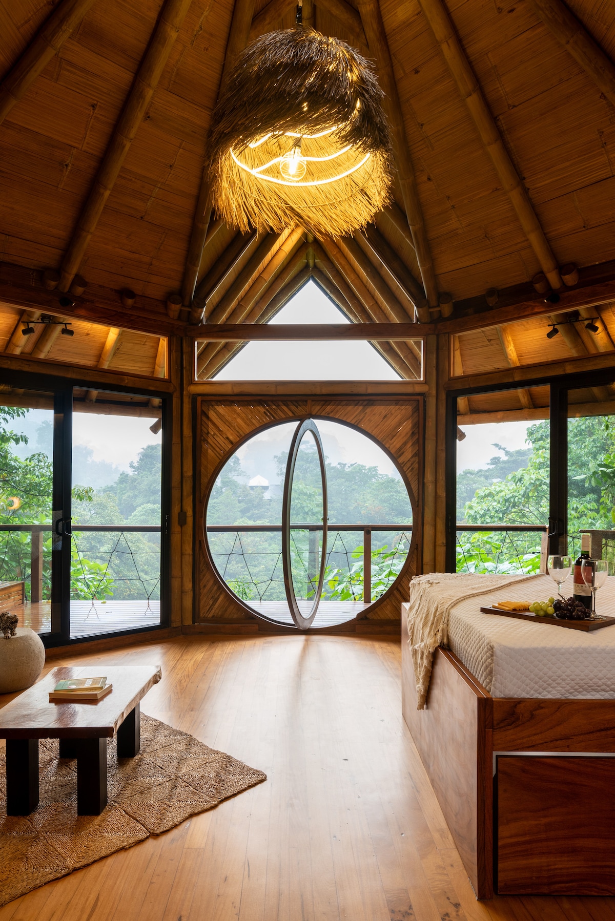 A circular window frames a lush green landscape, providing natural light to a spacious room with wooden accents. A low coffee table rests on a woven rug, and a bottle of wine is placed beside two glasses on the inviting bed.