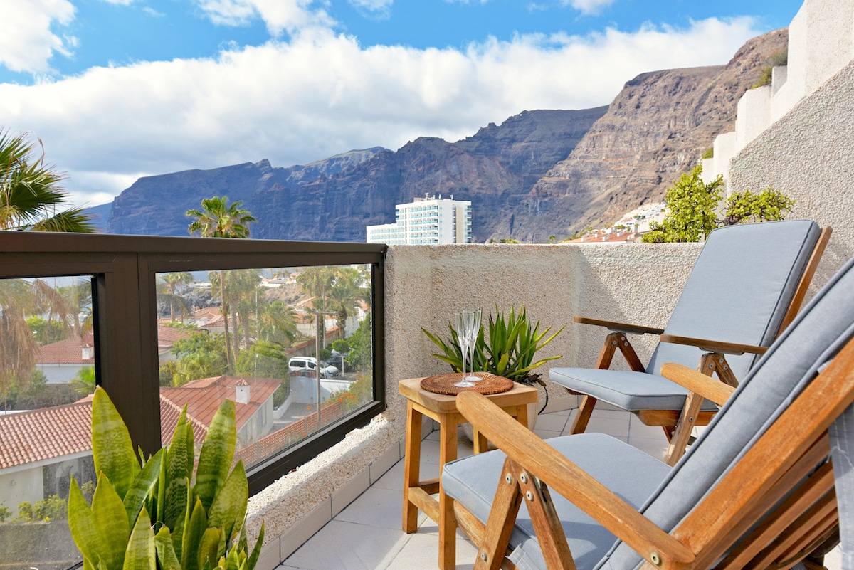 A spacious terrace features two comfortable chairs positioned for relaxation, with a small table nearby. Lush greenery is included in the decor. The backdrop showcases majestic mountains and a clear sky, providing a serene environment for outdoor enjoyment.
