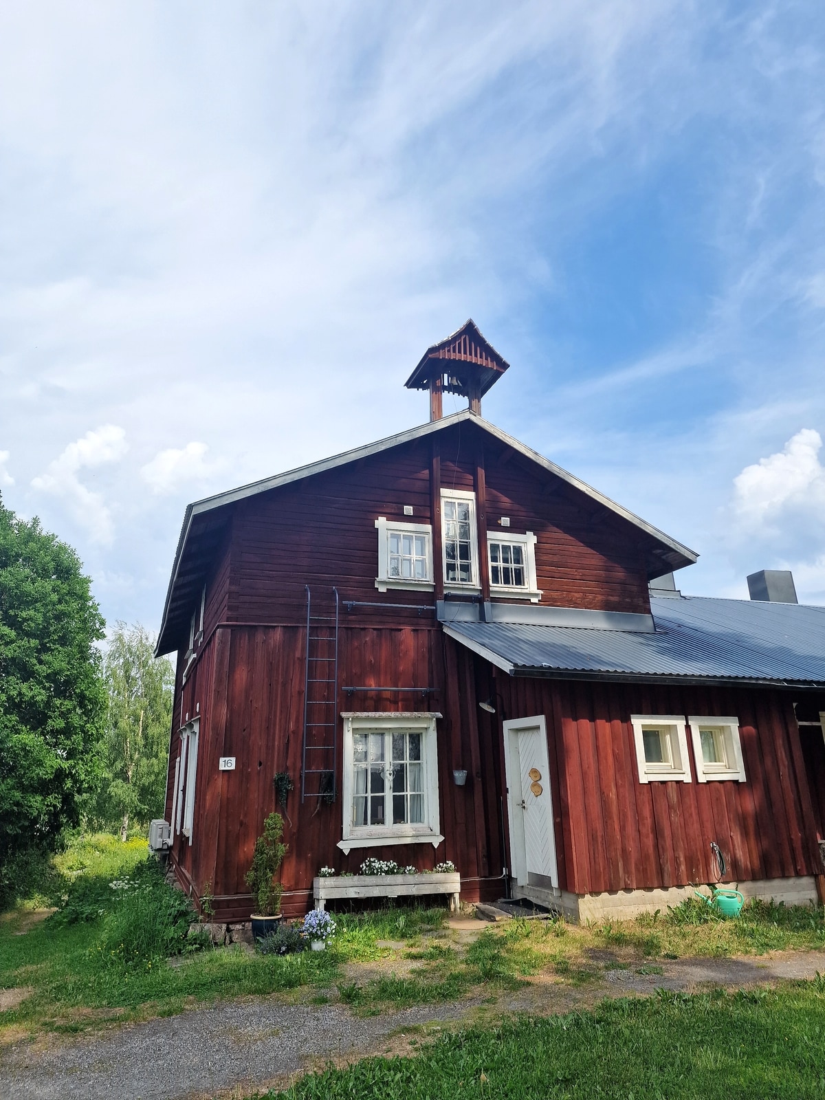 The exterior of a rustic two-story wooden villa is showcased, featuring a deep red facade and white-framed windows. A ladder leans against the side, and a small decorative structure sits atop the roof. Lush greenery surrounds the building, adding to its charm.