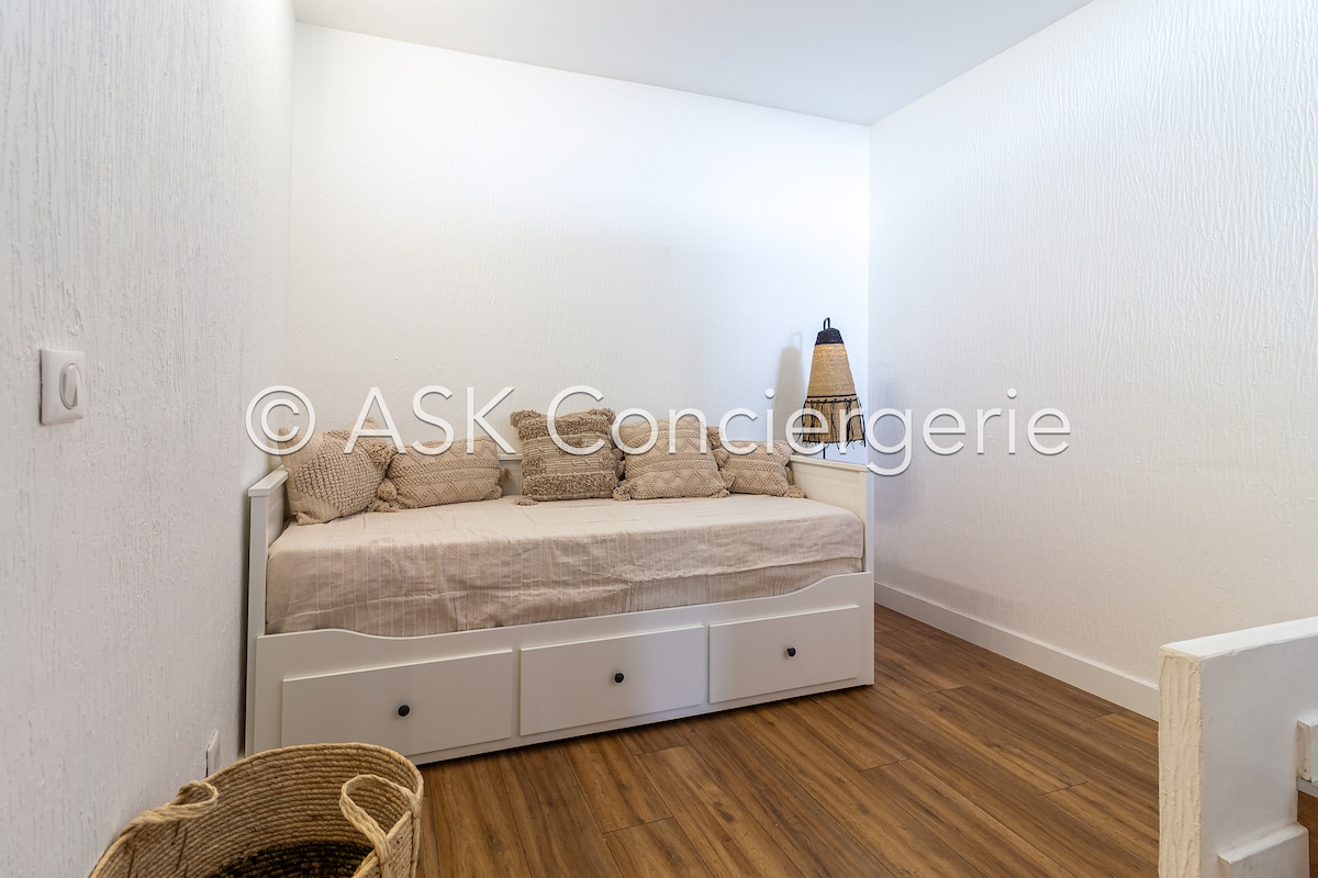 A light and airy room features a daybed with soft cushions against a textured white wall. Warm wooden flooring complements the minimalistic decor, with a woven basket placed nearby. A modern lamp provides additional lighting, enhancing the overall simplicity of the space.