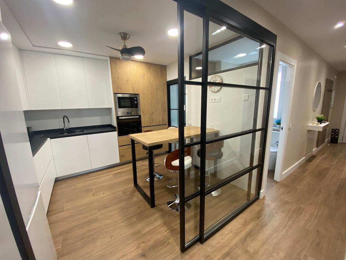 A modern kitchen area is featured, showcasing a sleek design with white cabinets and wood accents. A glass-enclosed dining space is visible, containing a wooden table with circular stools. The area is well-lit with recessed lighting, enhancing the contemporary atmosphere.