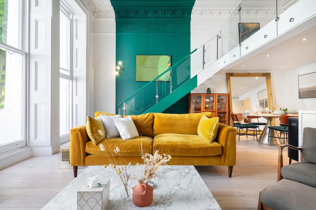 A spacious living area features a vibrant yellow sofa adorned with textured cushions, complemented by a marble coffee table. Large windows invite natural light, highlighting the intricate ceiling details and a colorful accent wall. A dining area and staircase are visible in the background.