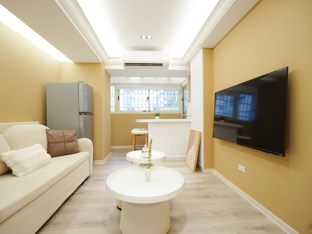 A bright living area features a comfortable light-colored sofa paired with a neutral accent pillow. Two white coffee tables are positioned in front, and a large flat-screen TV is mounted on the wall. Natural light filters through the window, enhancing the warm tones of the space.