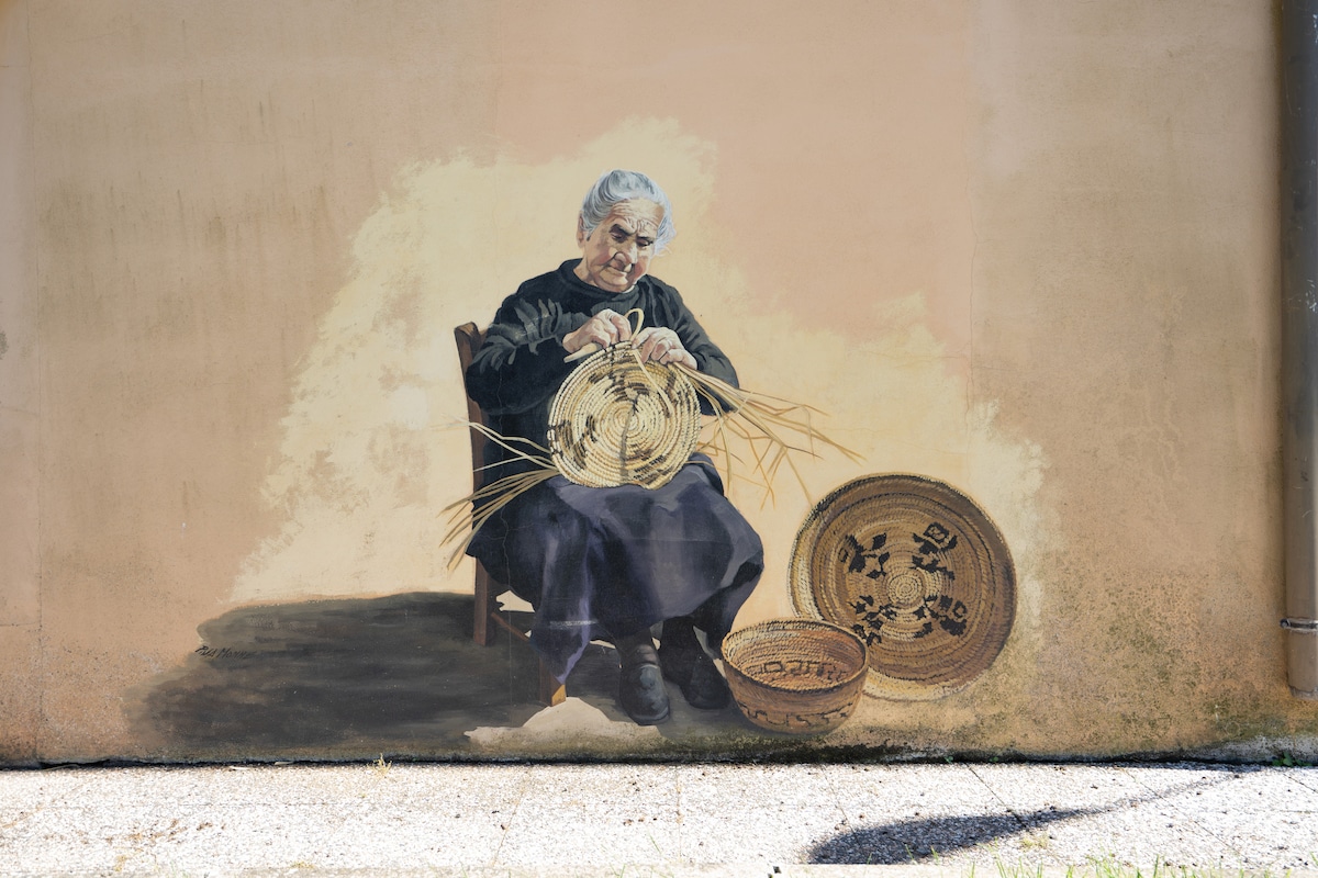 A mural depicts a seated figure crafting a woven basket, surrounded by finished baskets on the ground. The artwork is set against a textured, muted wall that enhances the scene, showcasing the artistry and tradition of basket weaving.