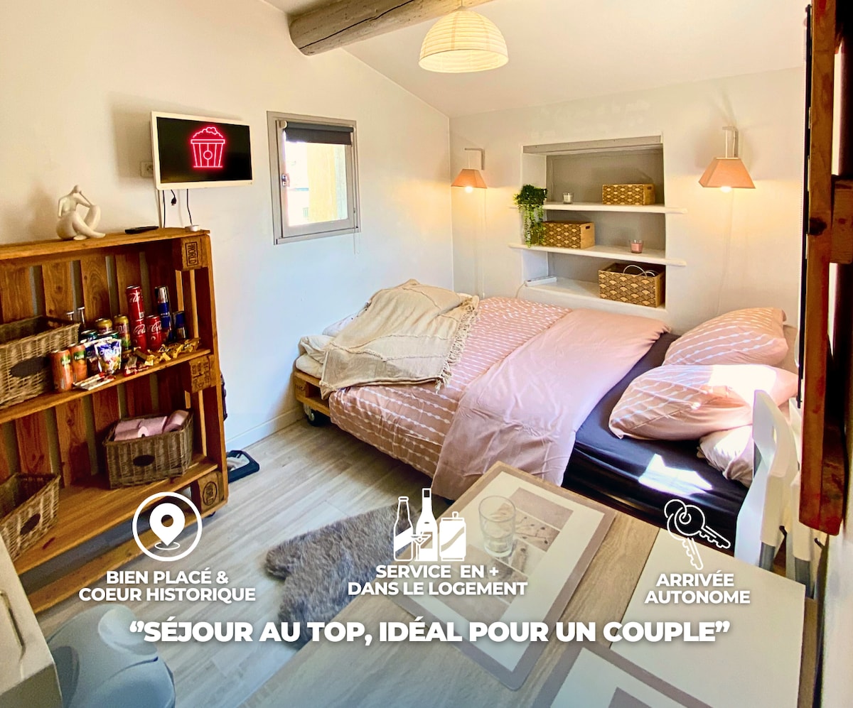 A cozy studio is presented with soft lighting and a minimalist design. A spacious bed, dressed in light pink linens, occupies the center of the room. Shelves hold a selection of snacks and beverages, enhancing the inviting atmosphere. A small window allows natural light to enter.