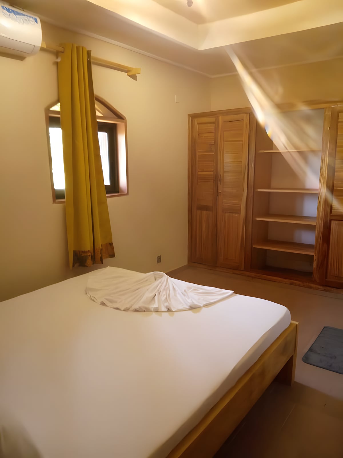 A comfortable bedroom features a spacious bed with a neatly arranged blanket. Natural light enters through a small window adorned with yellow curtains. Wooden wardrobes provide ample storage space, while minimalist decor emphasizes a serene atmosphere.