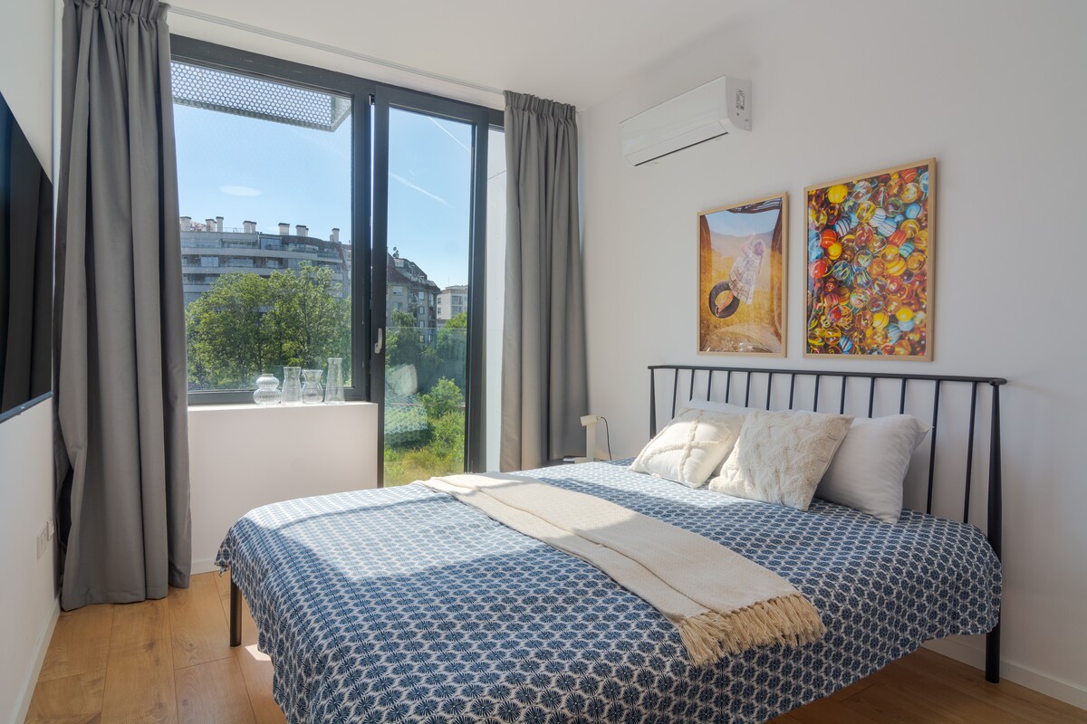 A modern bedroom features a spacious bed with a blue-patterned duvet and a light-colored throw. Two colorful pieces of wall art are displayed above the bed. Natural light fills the room through a large window with sheer curtains, offering a view of outdoor greenery.