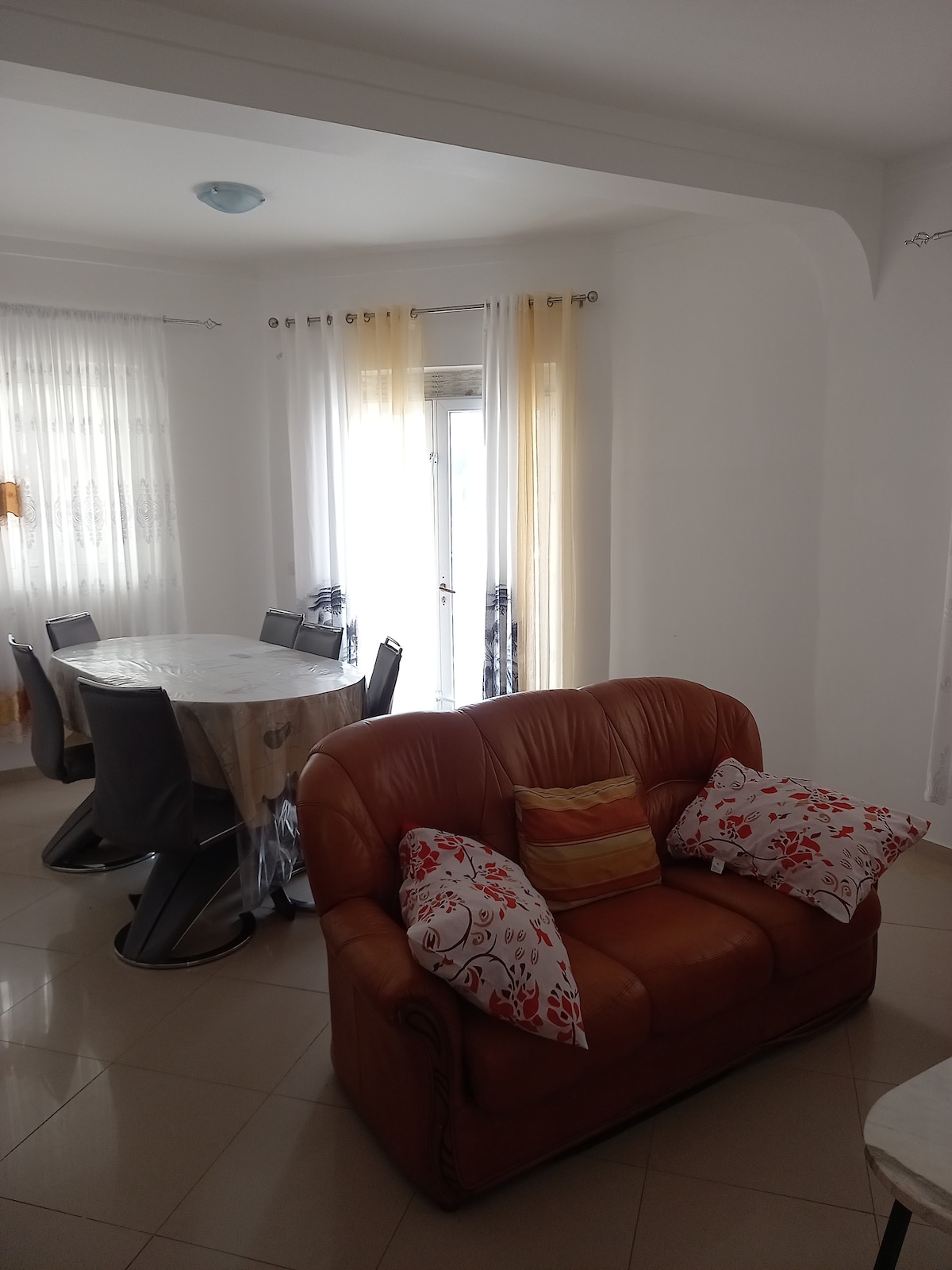 A living area is highlighted by a brown leather sofa adorned with patterned cushions. A round dining table is surrounded by sleek, modern chairs, and large windows allow natural light to brighten the space, complemented by sheer curtains.