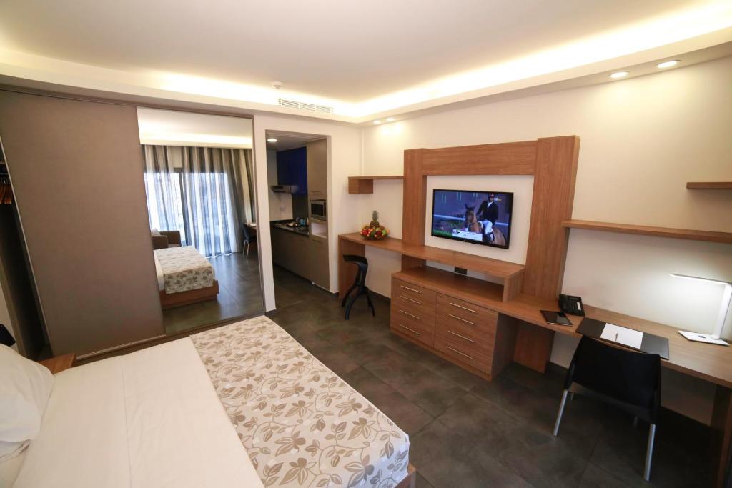 The studio room features a king-sized bed with a neutral-colored blanket. A flat-screen TV is mounted on a wooden shelf across from a work desk and chair. The kitchenette is visible in the background, and large windows provide natural light.