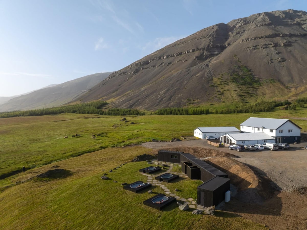 An aerial view captures the guesthouse and its surroundings, featuring multiple hot tubs nestled against a grassy field. A mountain rises majestically in the background, complementing the peaceful, open landscape. The guesthouse structure presents a modern design with large windows and a welcoming appearance.
