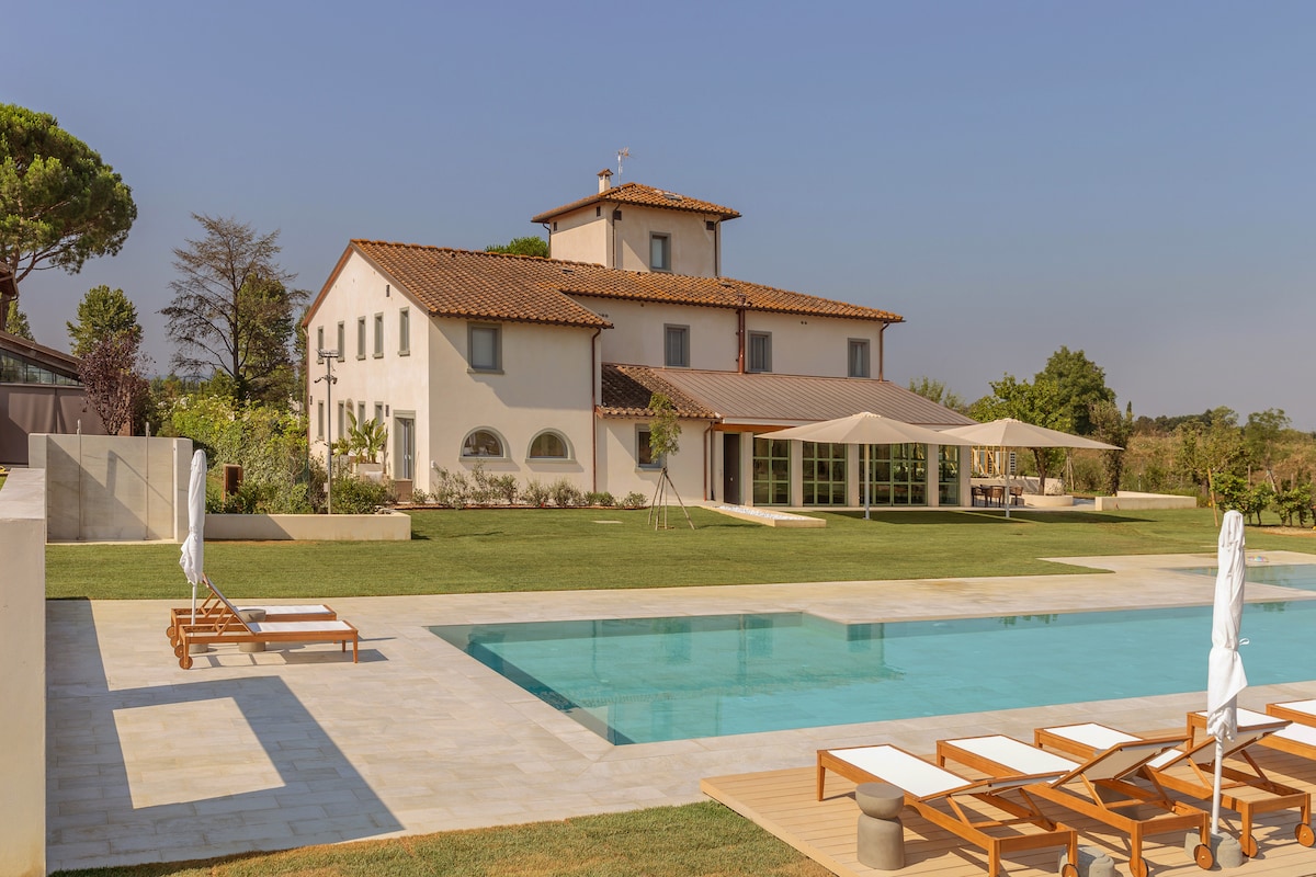 A modern villa with a traditional design is set against a clear sky. The property features a spacious pool area, an expansive lawn, and a covered terrace. Sun loungers are arranged alongside the pool, inviting relaxation amidst the serene landscape.
