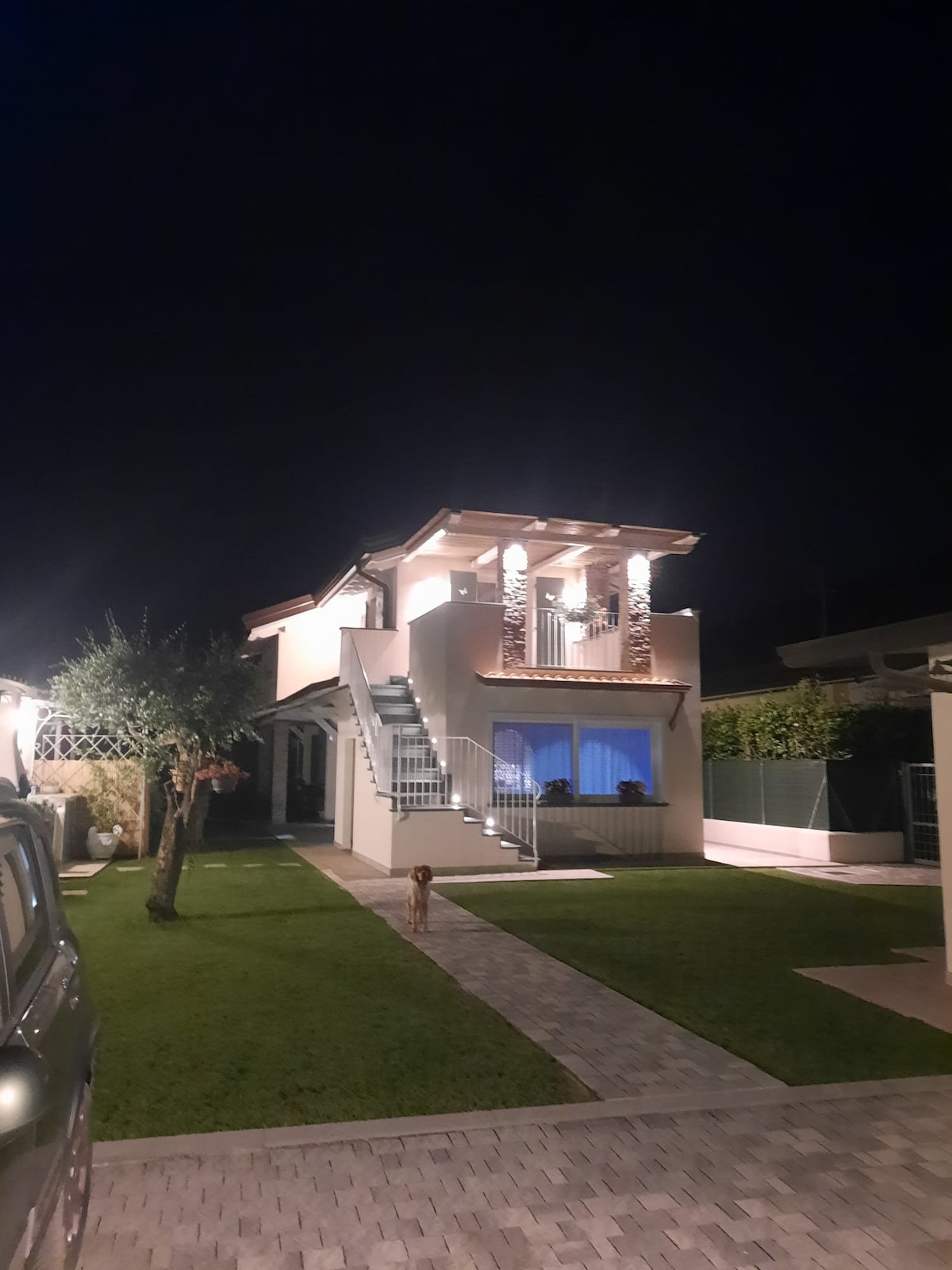 The two-story house is illuminated at night, showcasing its architectural features. A staircase leads to a covered balcony, where outdoor seating is visible. The surrounding area includes well-maintained grass and pathway, enhancing the home's inviting atmosphere.