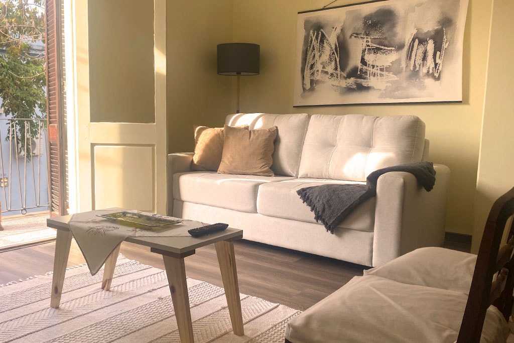 A light-filled living area features a neutral-colored sofa adorned with soft cushions. A wooden coffee table sits in front, accompanied by a cozy throw. Natural light filters in through an open door, illuminating a piece of wall art and enhancing the relaxing ambiance.