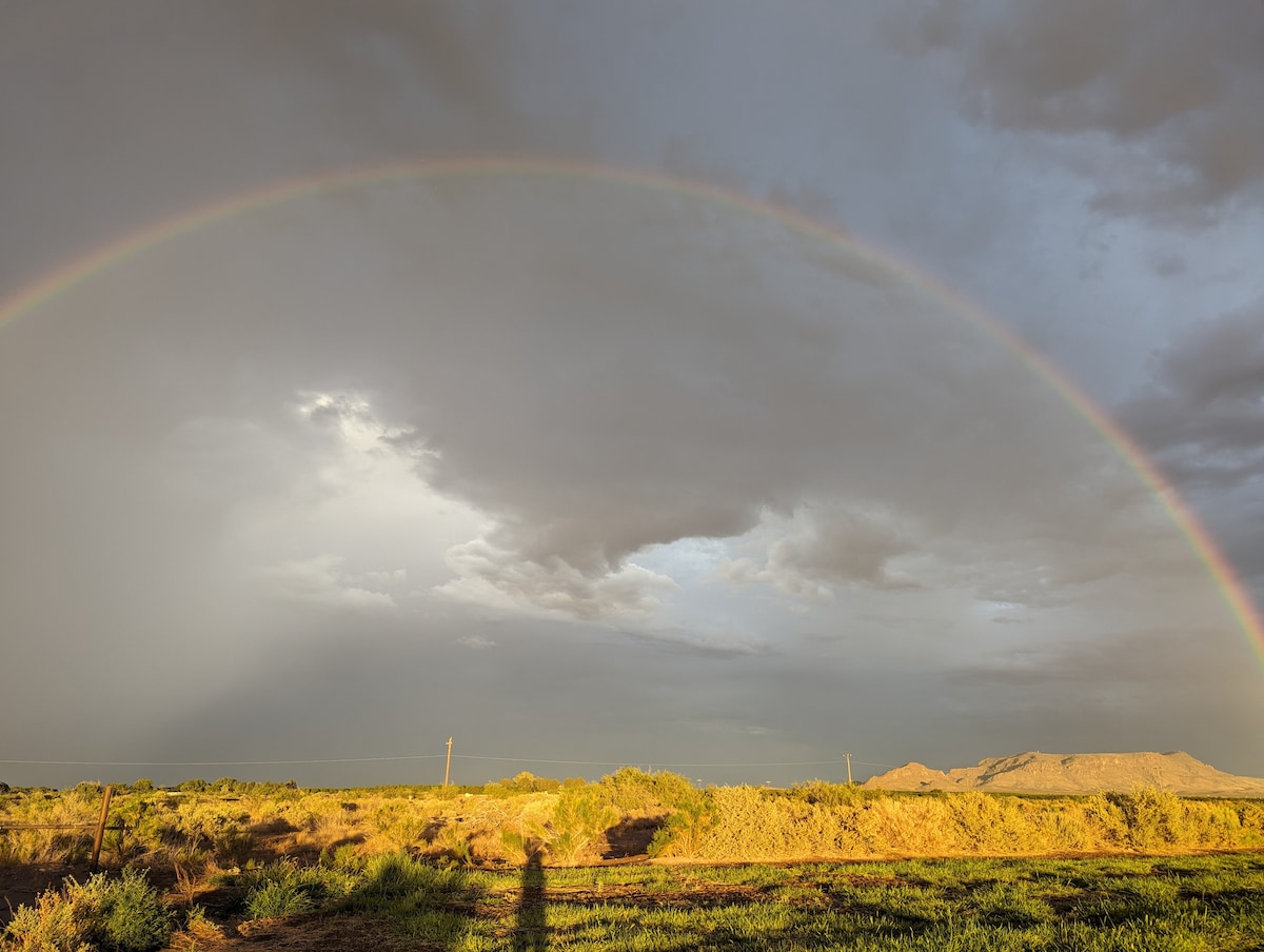 A broad rainbow arches across a cloudy sky, casting vibrant colors over a landscape of open fields and distant mountains. The natural scenery showcases various shades of green and brown, with textured clouds partially illuminated by sunlight.