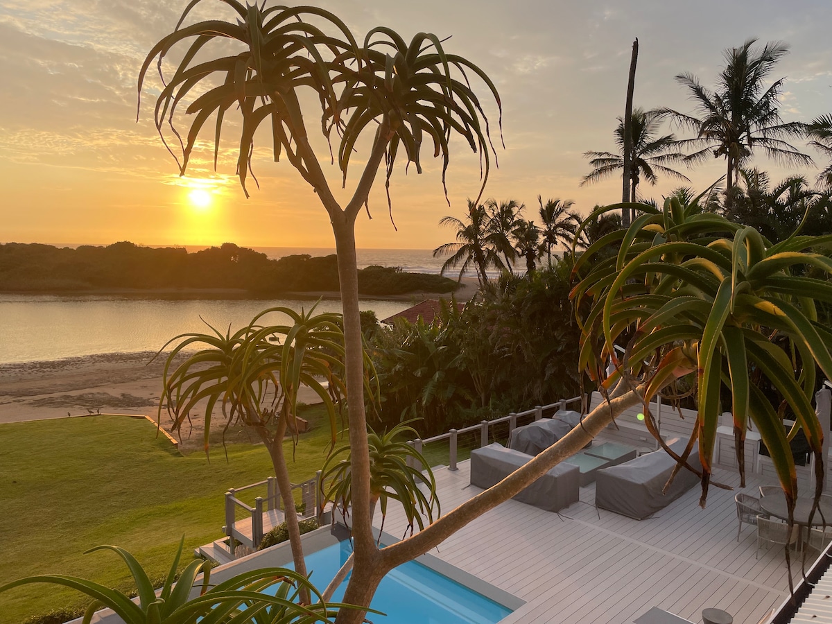 A serene sunset is captured over the lagoon, with palm trees framing the scene. The outdoor area features a well-maintained wooden deck, a swimming pool, and comfortable seating arrangements. The beach and lush greenery are visible in the background, enhancing the tranquil ambiance.