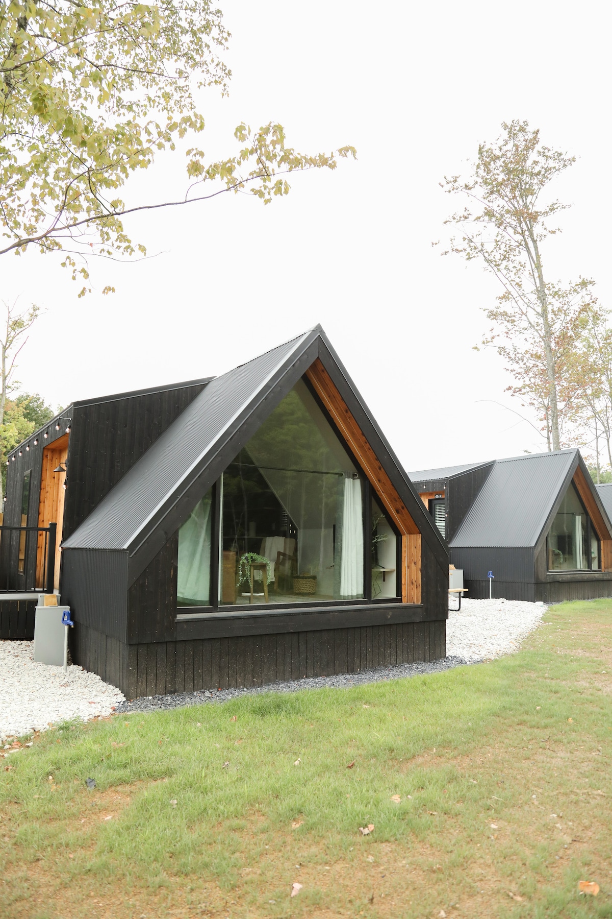 The exterior of a modern A-frame tiny home features a distinctive triangular roof and large glass windows. A grassy area surrounds the home, leading to a charming outdoor space with seating. The design offers a seamless connection to the surrounding nature.