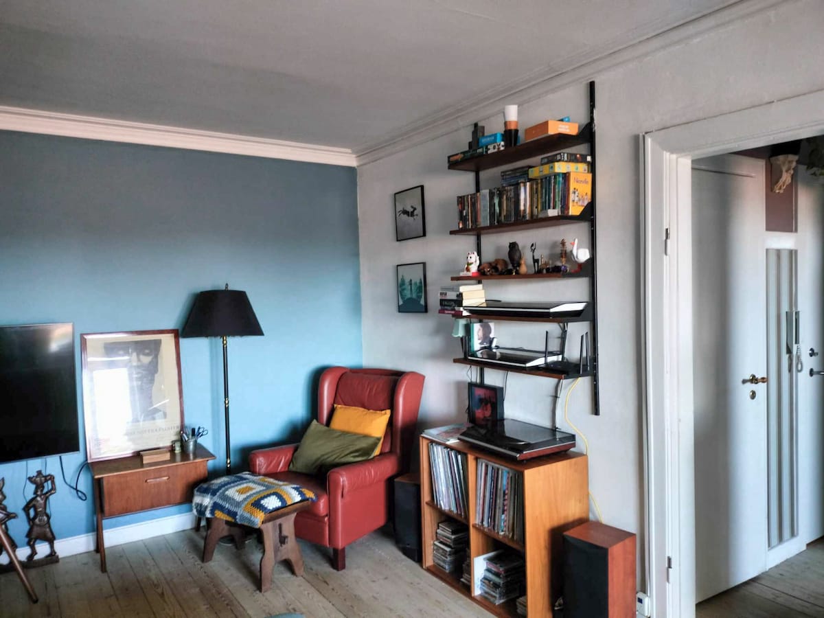 A cozy corner of the living room is shown, featuring a red armchair with a colorful throw blanket draped over the side. A small lamp stands nearby, illuminating the space. Open shelving displays books and decorative items, while a television sits opposite the seating area.