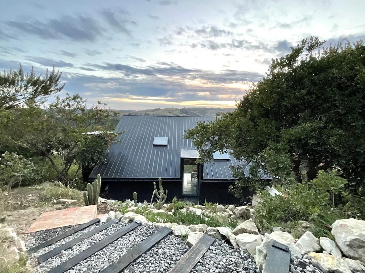 A modern tiny house with a sleek black roof is surrounded by native vegetation. The pathway is lined with stones and gravel, leading towards the home's entrance. The backdrop features a scenic view of the landscape under a soft, cloudy sky during sunset.