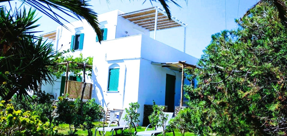 The exterior of a white two-story building is surrounded by lush greenery and flowering plants. Windows with green shutters add color to the façade, while a rooftop terrace provides additional outdoor space. Lounge chairs are positioned on the grassy area for relaxation.