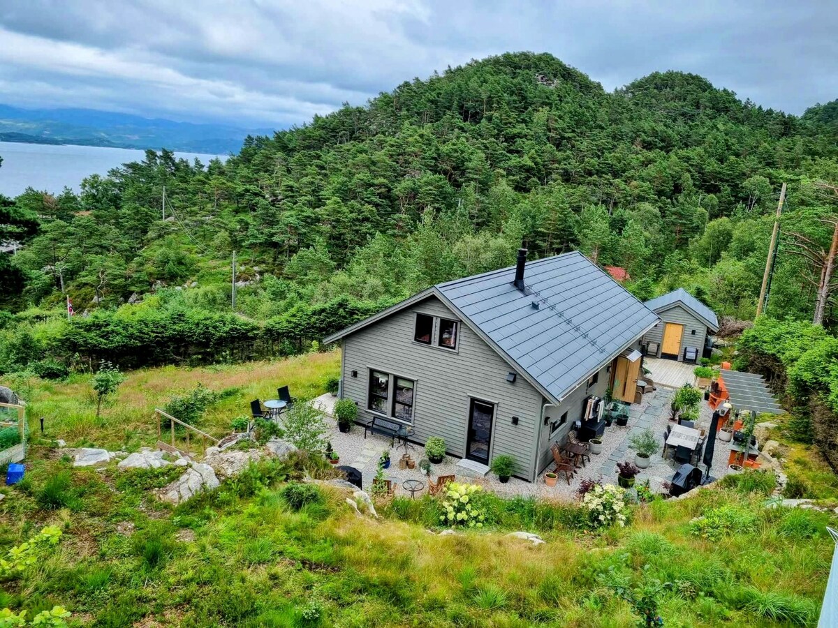 A modern cabin is nestled among lush greenery, featuring a sloped roof and large windows. Several outdoor seating areas are visible, surrounded by well-maintained garden beds. The serene landscape is complemented by views of nearby hills and water, promoting a tranquil retreat.