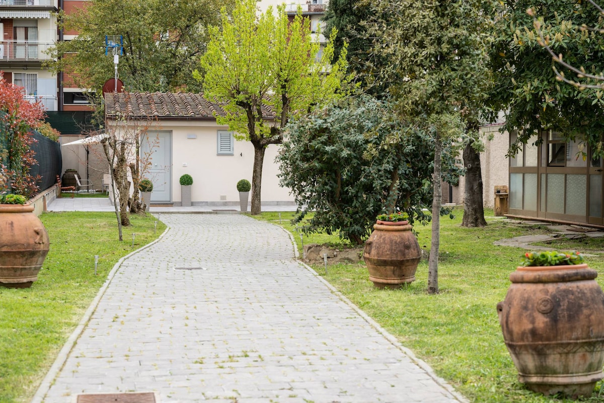 A pathway paved with stones leads through a landscaped garden, lined with decorative terracotta pots. Lush greenery and small trees are visible, creating a serene outdoor environment. A building with a light-colored exterior stands in the background, partially obscured by foliage.