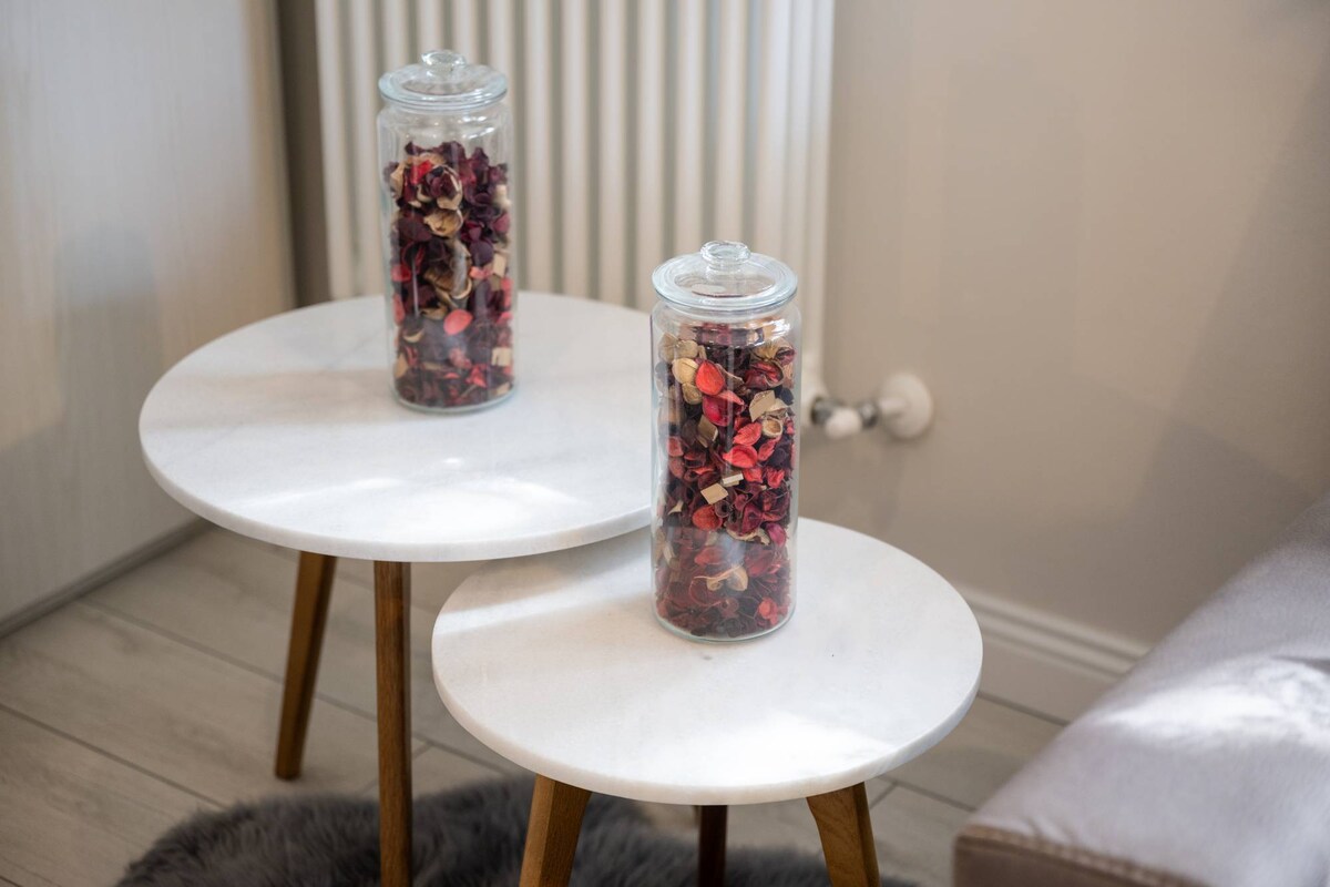 Two round white marble side tables are positioned near a soft, neutral-colored sofa. Each table is topped with a glass jar filled with colorful dried petals, adding a touch of natural decor to the space. A radiator is visible in the background.