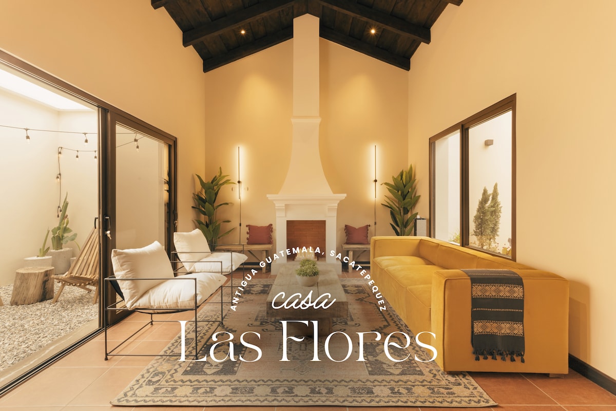 A spacious living area features a modern fireplace, white walls, and a combination of comfortable seating. Large glass doors allow natural light to fill the space, with potted plants adding a touch of greenery. The floor is tiled, enhancing the room's clean aesthetic.