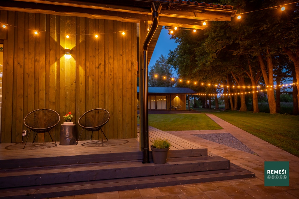 A cozy outdoor seating area is illuminated by warm string lights, enhancing the evening ambiance. Two comfortable chairs are positioned near a small table on a wooden deck, with a well-maintained grassy area extending into the distance.