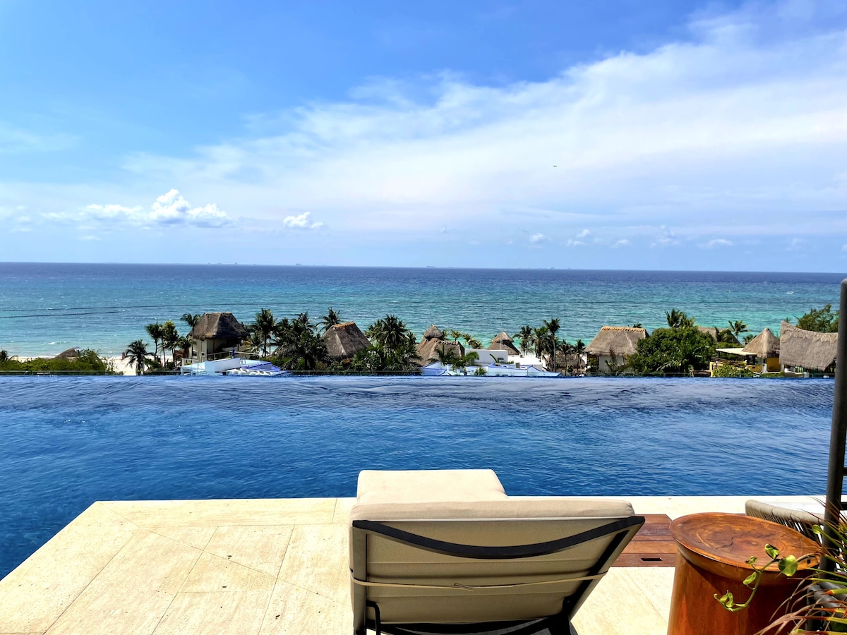 An infinity pool overlooks the serene ocean, blending seamlessly with the horizon. Comfortable lounge chairs are placed nearby, providing a relaxing spot to enjoy the view. Lush greenery and thatched-roof structures are visible along the coastline, enhancing the tranquil atmosphere.