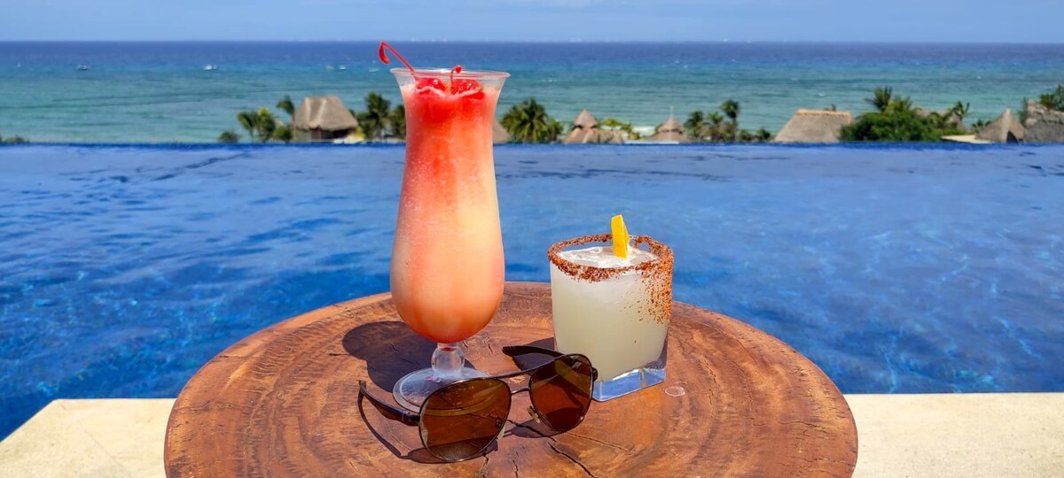 Two refreshing cocktails are presented on a wooden table with an ocean view. A vibrant, layered drink features a cherry on top, while a salted rimmed glass contains a light-colored beverage with a lemon slice. The infinity pool stretches toward the sea, enhancing the tranquil atmosphere.