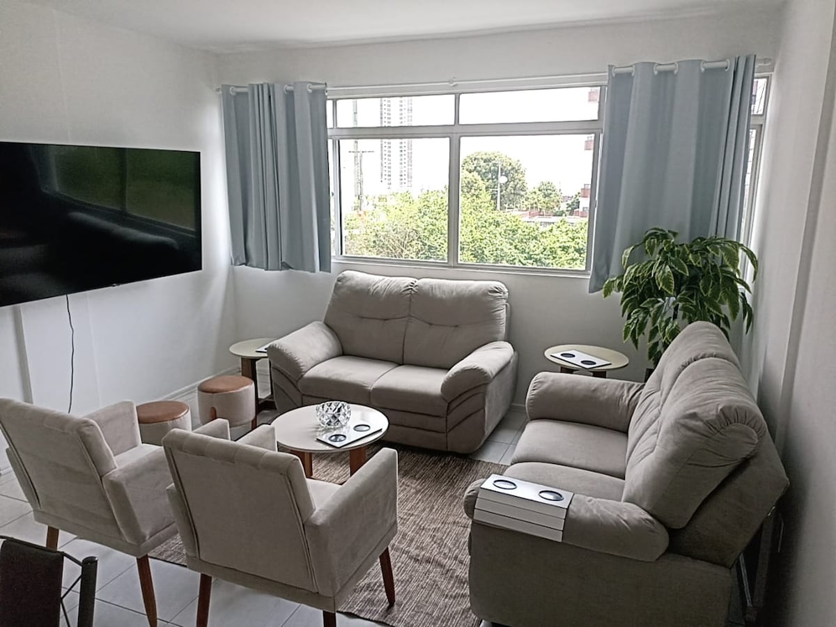 A comfortable living area is enhanced by large windows that allow natural light to fill the space. The seating arrangements include a sofa, armchairs, and small tables, with a television mounted on the wall and greenery visible outside.