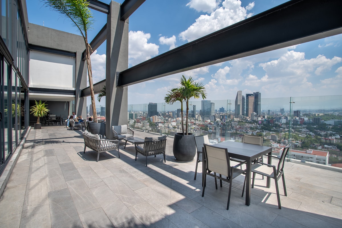 An expansive rooftop terrace offers stunning city views, featuring modern seating arrangements and a dining table surrounded by greenery. The space is adorned with palm plants, and large glass barriers provide clear sightlines of the skyline, enhancing the outdoor experience.
