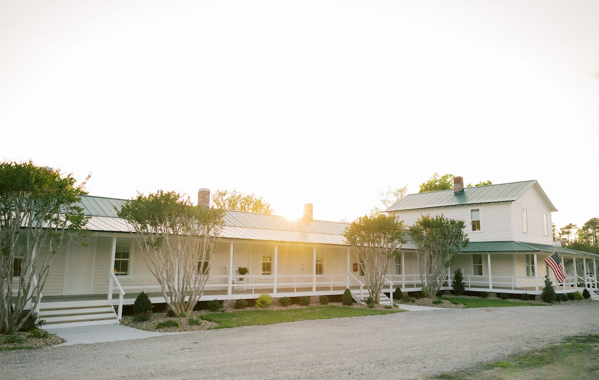 The exterior of The Bunk House is showcased, featuring a two-story building with a welcoming porch and vibrant greenery along the path. A soft glow from the setting sun illuminates the structure, highlighting the charm of its historic design.