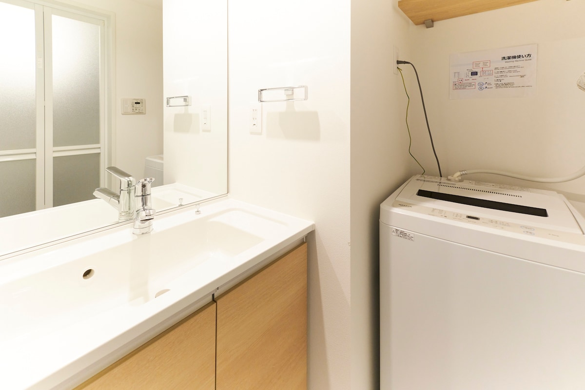 A modern bathroom features a sleek countertop with an integrated sink and a large mirror above it. Next to the sink, a washing machine is positioned under a wooden shelf, providing convenience for guests. Soft natural light illuminates the space.