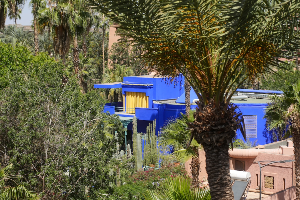 The image features a vibrant blue building surrounded by lush greenery, including palm trees and various plants. Yellow curtains are visible, offering a glimpse of the interior, while the colorful structure stands out amidst its natural surroundings.