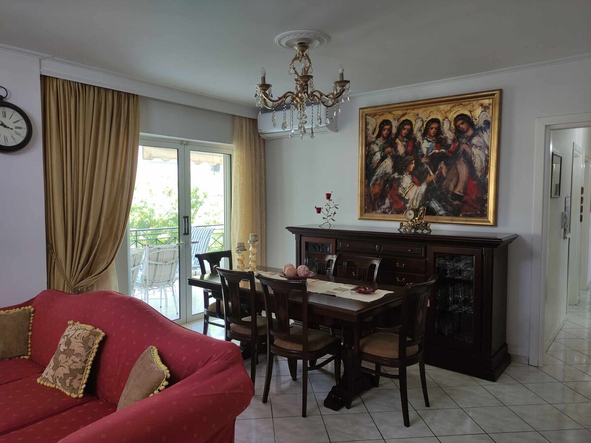 A spacious living and dining area is featured, with elegant furnishings including a mahogany dining table surrounded by wooden chairs. A vibrant artwork adorns the wall, while large windows allow natural light to fill the space, complemented by soft drapes and a chandelier overhead.