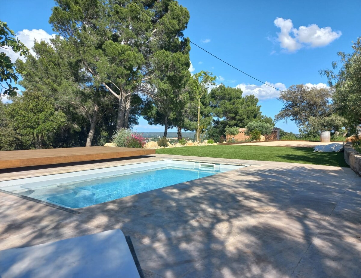 A serene outdoor space is highlighted by a crystal-clear swimming pool surrounded by lush greenery. Sunlight filters through the trees, casting gentle shadows on the patio area. The distant landscape is visible, providing a sense of tranquility and connection to nature.