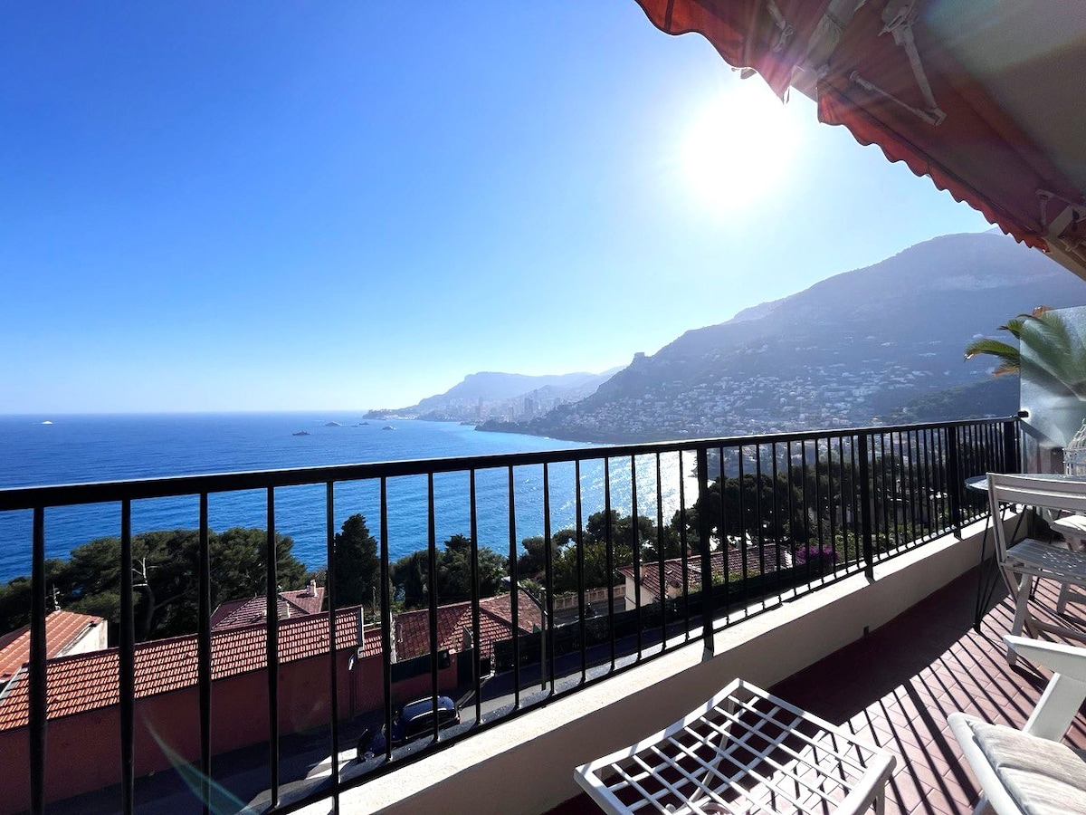 A panoramic view of the coastline and nearby hills is displayed from a terrace. The bright sun illuminates the scene, highlighting the tranquil blue of the sea and the lush greenery below. A railing delineates the terrace, with comfortable seating positioned to enjoy the vista.
