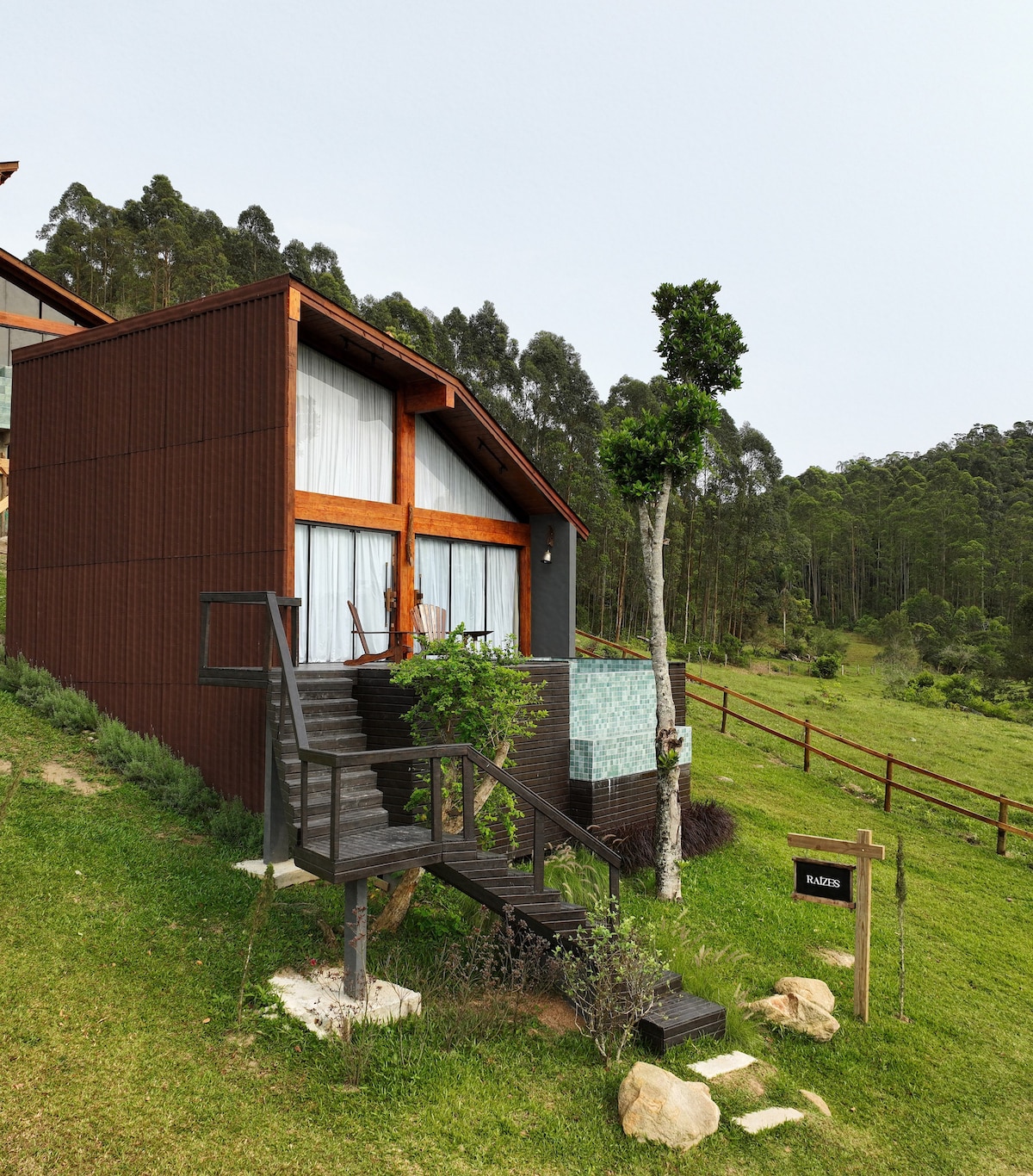 The exterior of the cabin is surrounded by lush greenery, showcasing a contemporary design with large windows. Steps lead up to the entrance, flanked by a small garden area. A sign indicating 'Raízes' is visible, complementing the natural landscape.