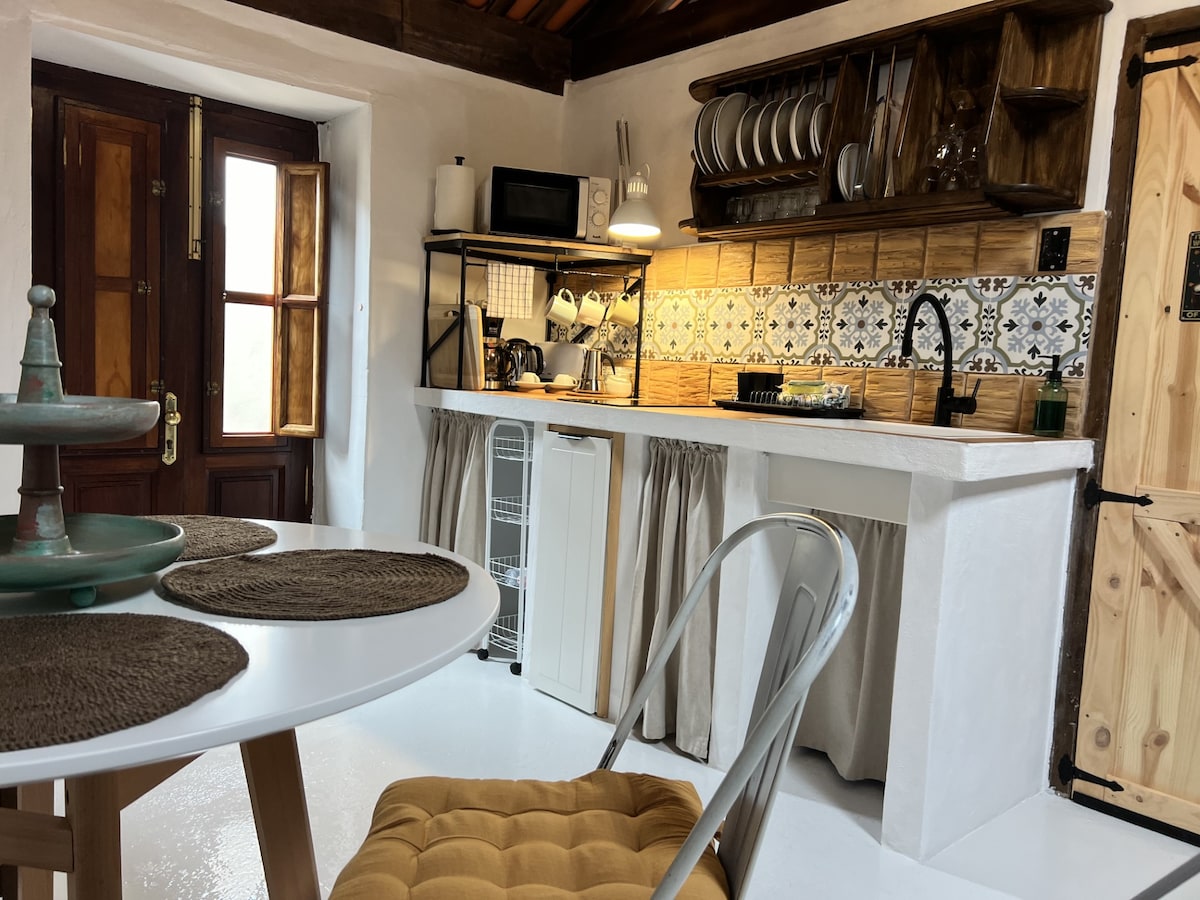 A charming kitchen area features open shelving displaying dishware and utensils. A modern sink and stove are situated against patterned tiles. A round dining table with textured place mats is complemented by a metal chair, enhancing the cozy and functional space.