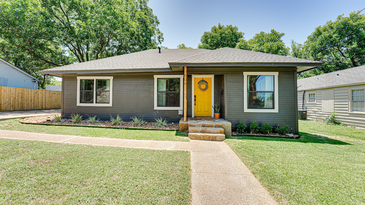 The exterior of the home features a welcoming entrance with a vibrant yellow door. Lush green grass surrounds the property, complemented by neatly arranged shrubs. The structure has multiple windows, allowing natural light to enhance its modern façade, while trees provide shade in the background.