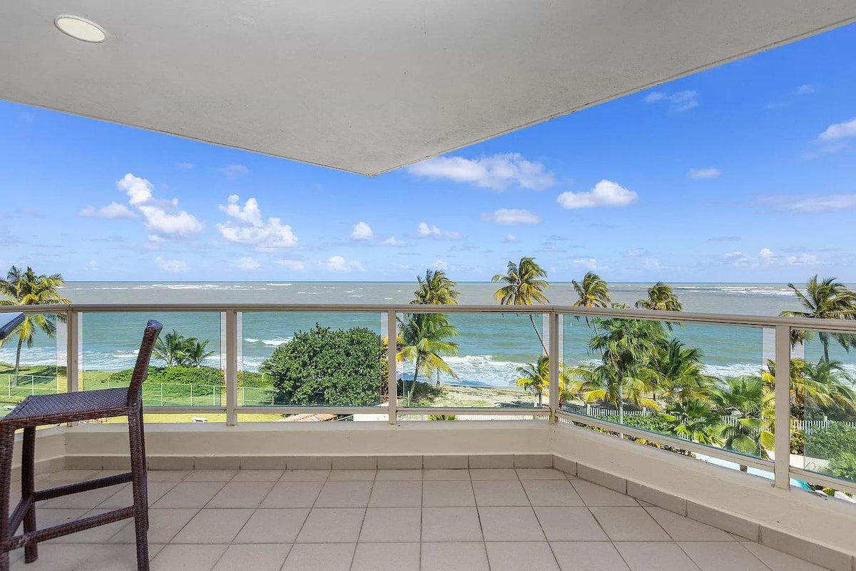 A private balcony is showcased, featuring a single woven chair. The view overlooks the ocean with gentle waves and lush palm trees lining the shore. Clear blue skies contrast with the greenery, creating a tranquil outdoor space for relaxation.