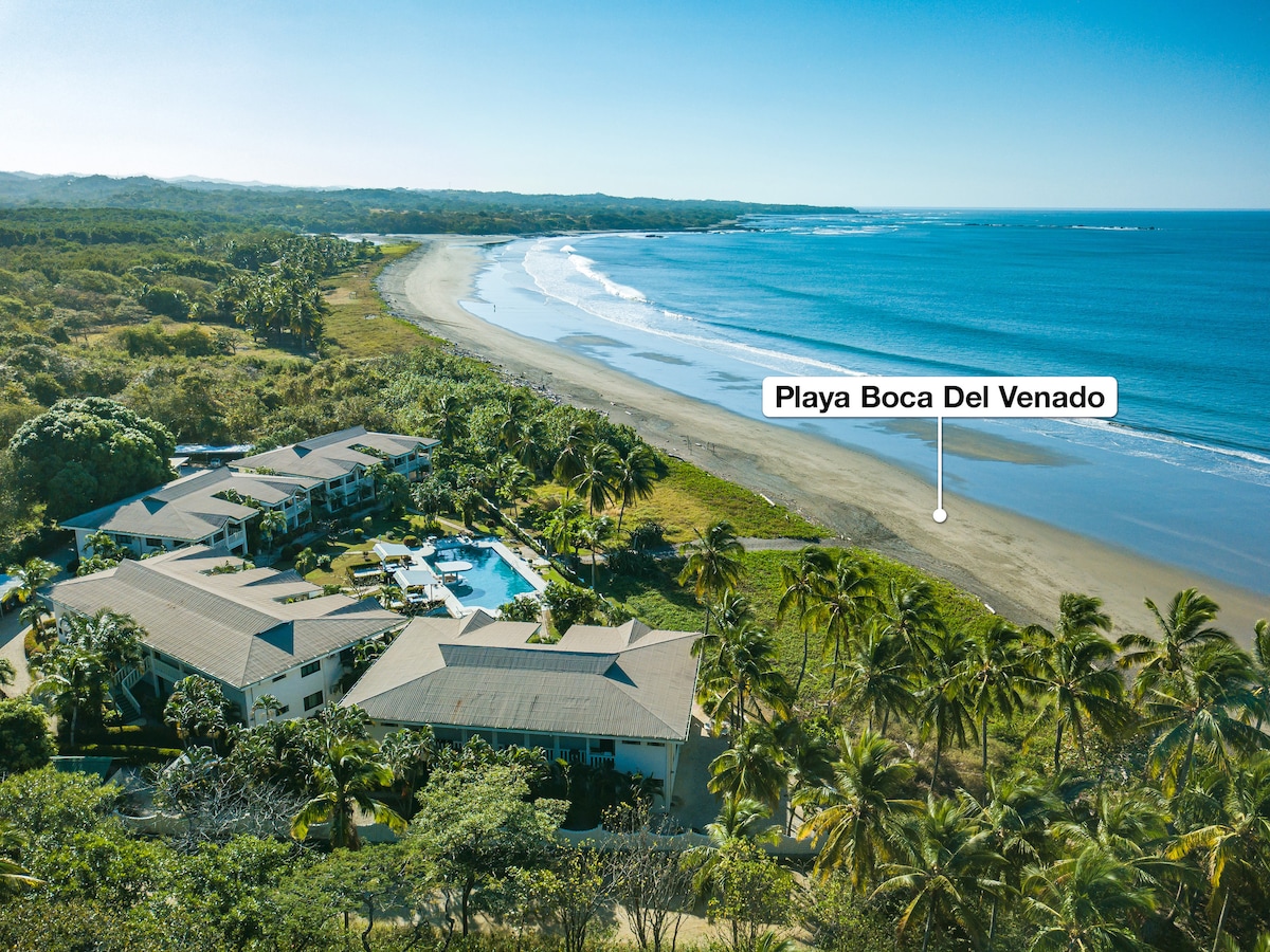 An aerial view captures the beachfront condominium nestled among lush greenery. A communal pool glistens in the center, surrounded by palm trees. The expansive sandy beach stretches along the coastline, with gentle waves lapping at the shore, offering a serene coastal landscape.