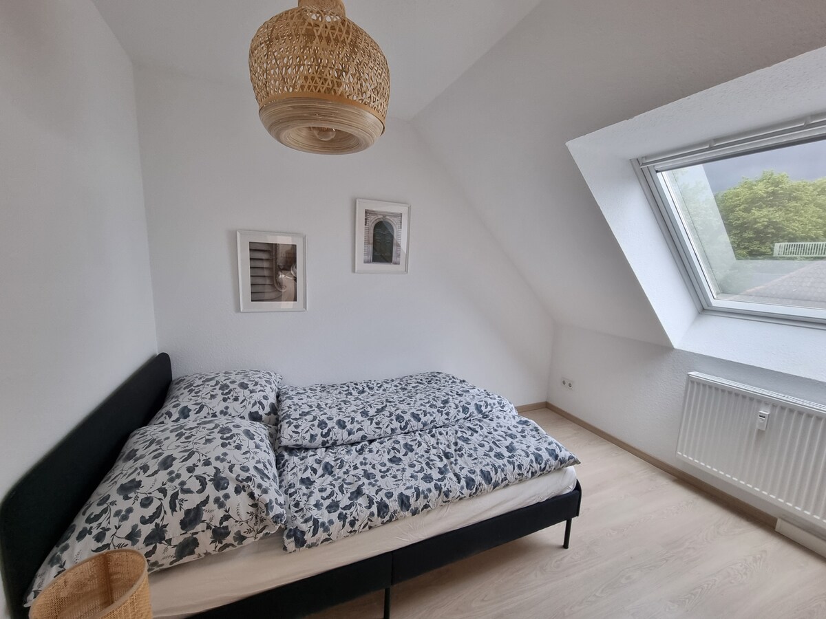 A bright bedroom features a spacious bed with patterned bedding, complemented by two framed prints on the walls. Natural light enters through a large angled window, illuminating the light-colored wooden floors and minimalist decor, contributing to a tranquil environment.