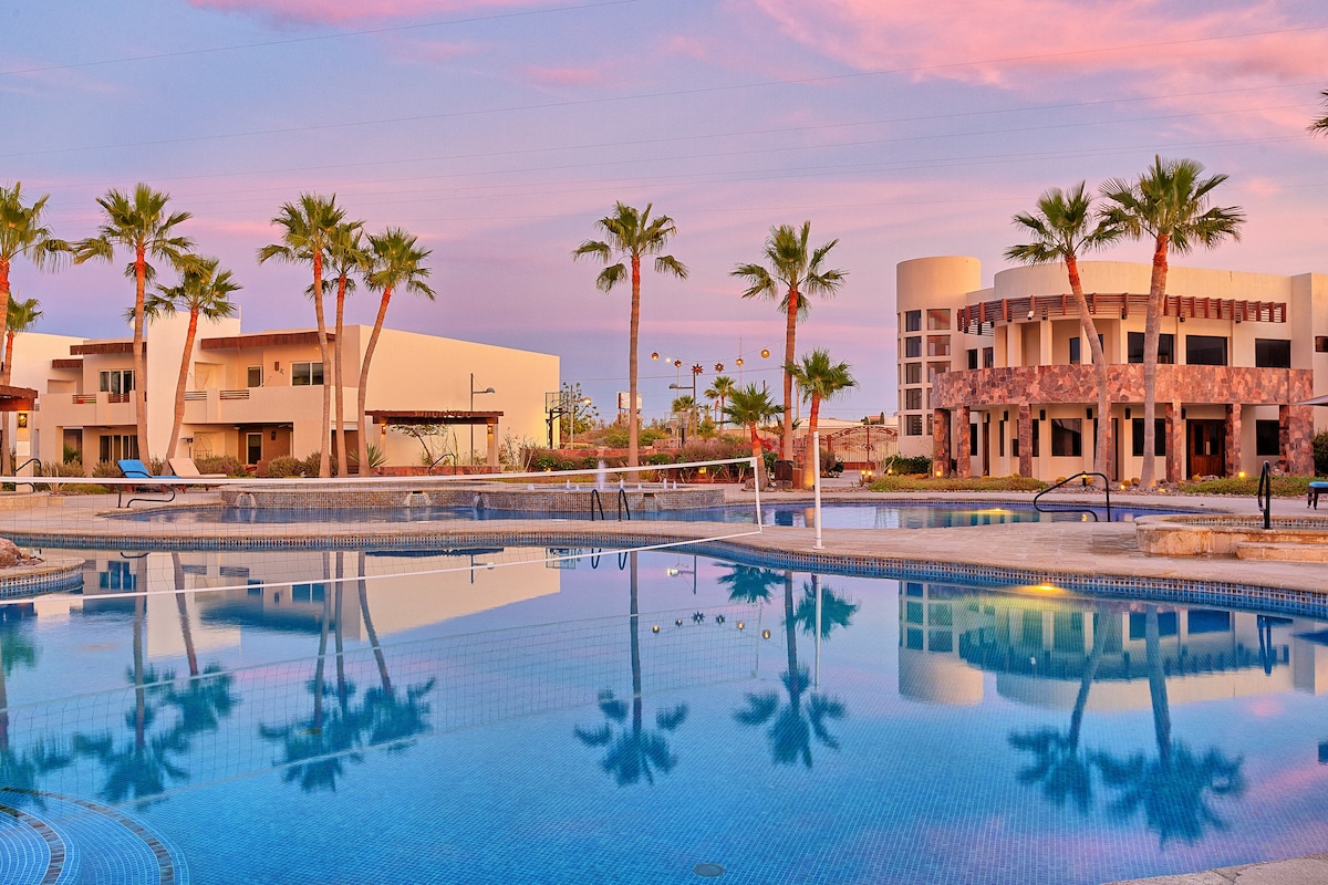 A large swimming pool reflects the colorful sky during sunset. Palm trees surround the area, and modern buildings are located in the background. The scene conveys a relaxed atmosphere, ideal for enjoying the outdoors.