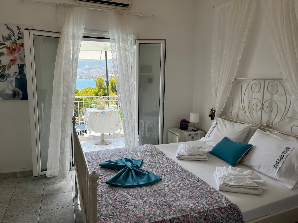 A bright and airy bedroom features a spacious bed adorned with white linens and turquoise accents. Floor-to-ceiling glass doors open to a balcony with views of the surrounding landscape. Light drapes softly frame the entrance, enhancing the connection with the outdoor space.