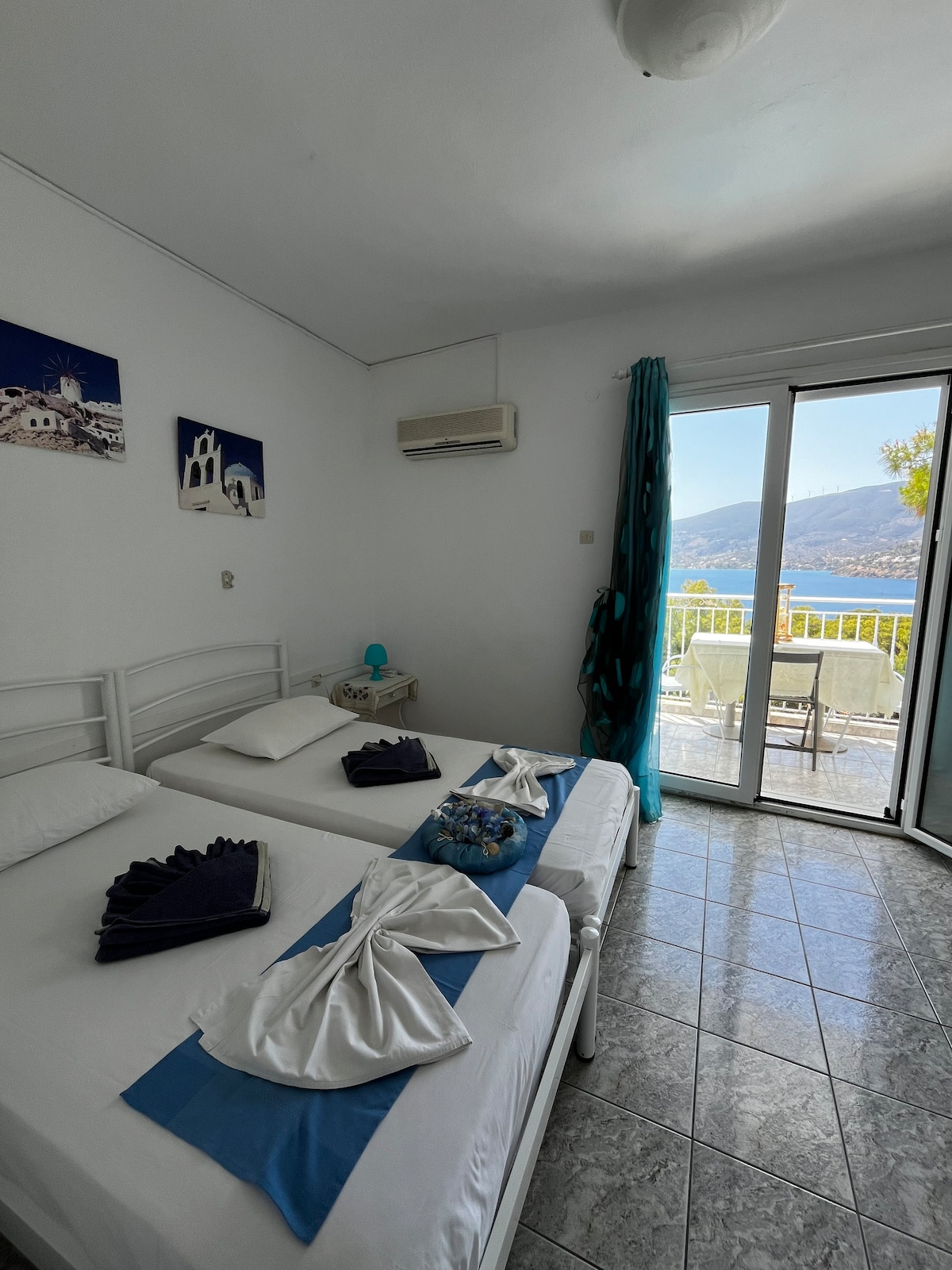 A bright and airy room contains two twin beds dressed with crisp linens, complemented by decorative towels. A sliding glass door provides access to a balcony with a scenic view. Blue accents from drapes and decor add a refreshing touch to the space.