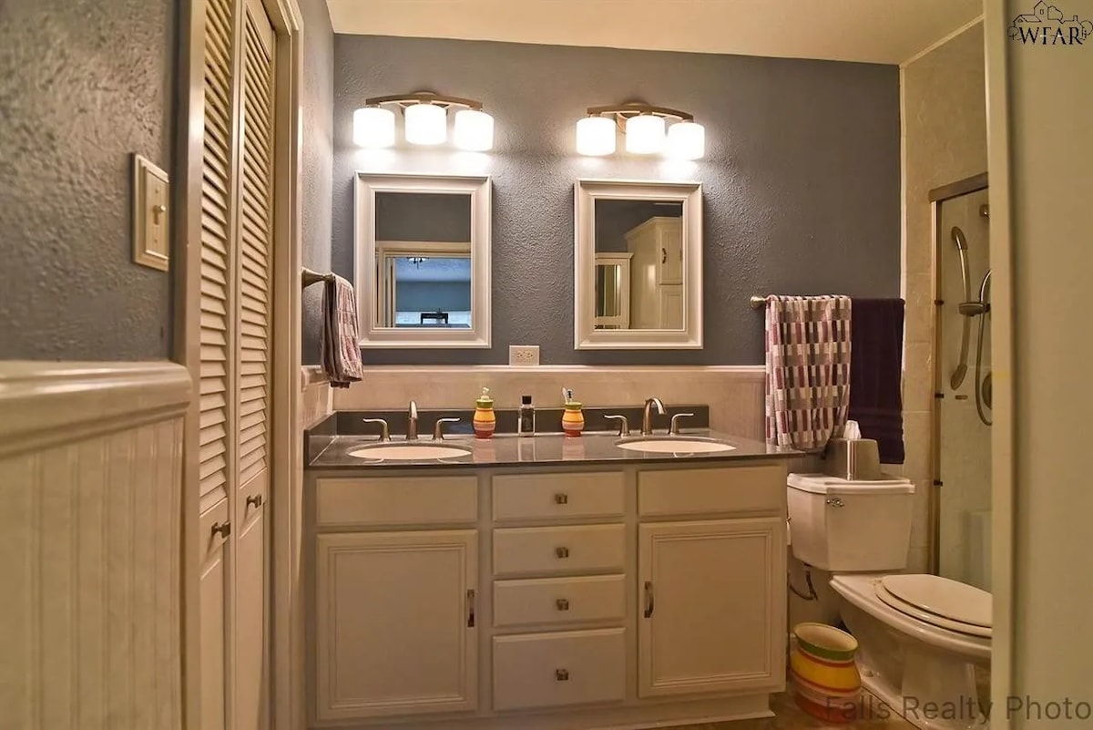 A well-lit bathroom features dual sinks with coordinated mirrors above each. Cabinets provide ample storage, and a shower area is visible in the corner. The walls are painted in a calming blue, complemented by a white wainscoting trim.