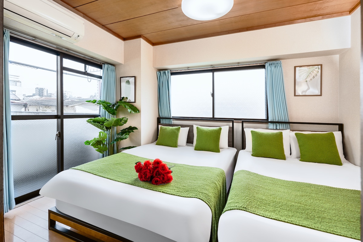A well-lit bedroom features two spacious beds covered with bright white linens and textured green throws. Pillows in complimentary green hues are arranged neatly. Natural light enters through large windows adorned with light blue curtains, and a small potted plant adds a touch of greenery.